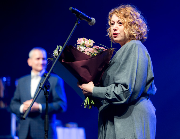 Na zdjęciu kobieta trzymająca kwiaty mówi coś do mikrofonu