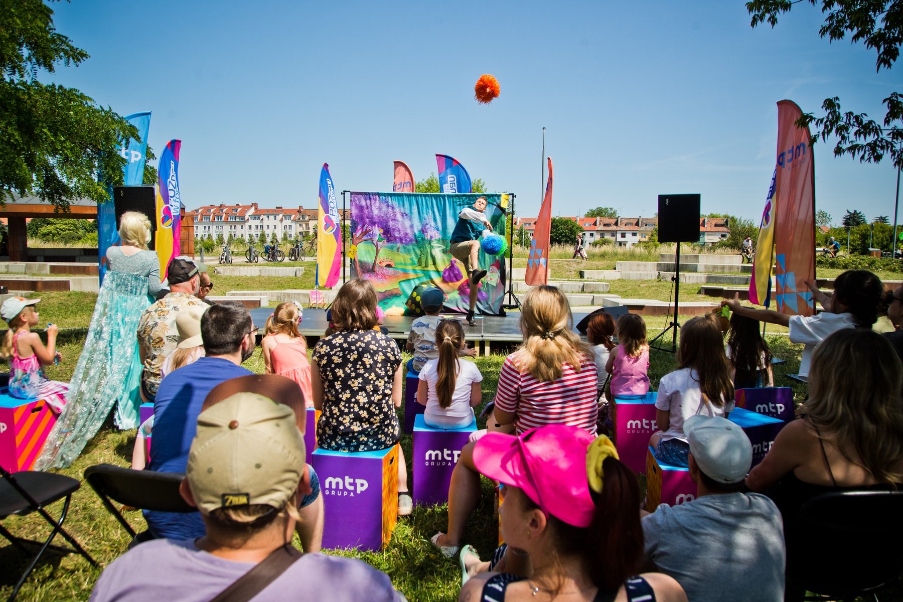 Na zdjęciu ludzie zebrani przed sceną na świezym powietrzu, na pierwszym planie widać gałęzie drzew