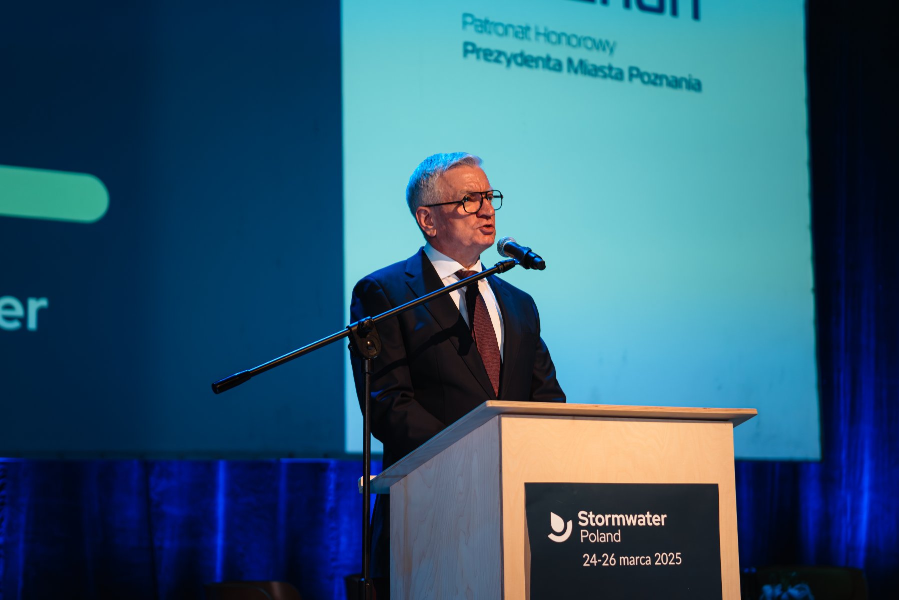 Na zdjęciu meżczyzna na scenie, w tle ekran, na nim napis: patronat honorowy Prezydenta Miasta Poznania