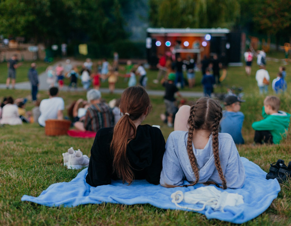 Na zdjęciu młodzi ludzie na trawie, na kocu, tyłem do obiektywu