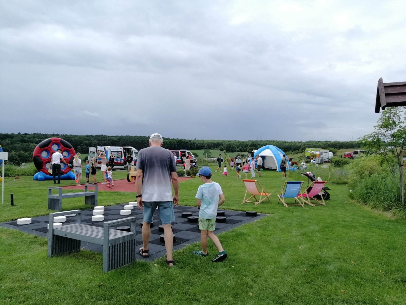 Na zdjęciu, na piewrszym planie widać wielką szachownicę do plenerowych warcabów. Dwie osoby - mężczyzna i dziecko - stoją przy szachownicy. W tle widoczne sa inne atrakcje piknikowe, leżaki, namioty i pojazd ratownictwa medycznego