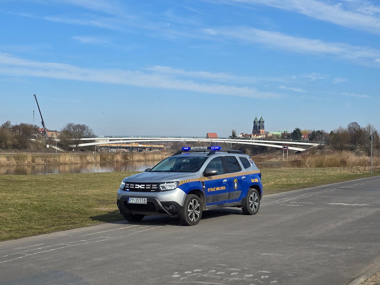 Na zdjęciu radiowóz straży miejskiej nad Wartą podczas patrolu. W tle nowe mosty Berdychowskie oraz Katedra. Ładny słoneczny dzień, zachęcający do spacerów.