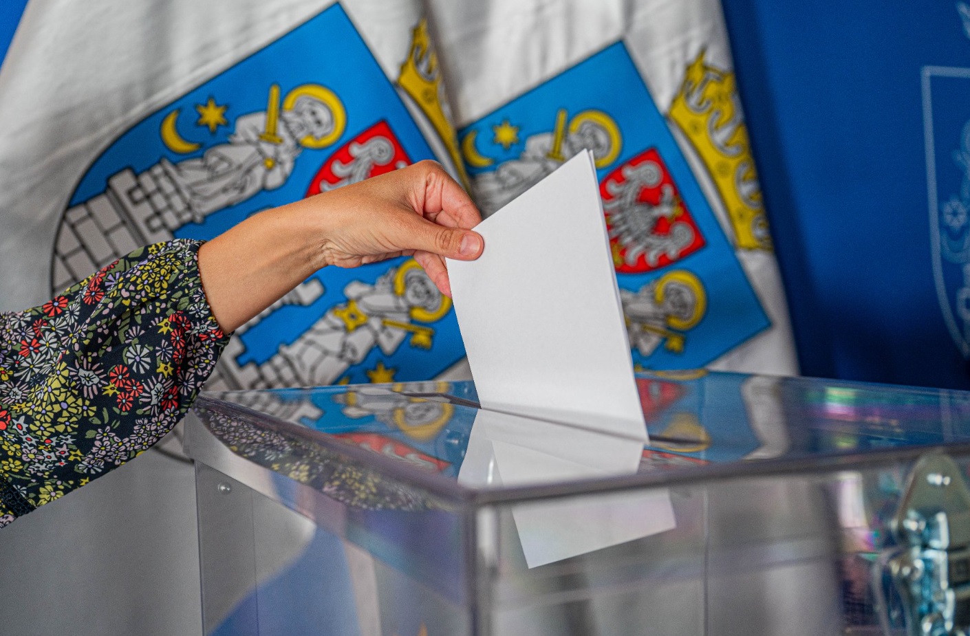 Na zdjęciu ręka wrzucająca głos do urny, w tle flagi z herbem Poznania