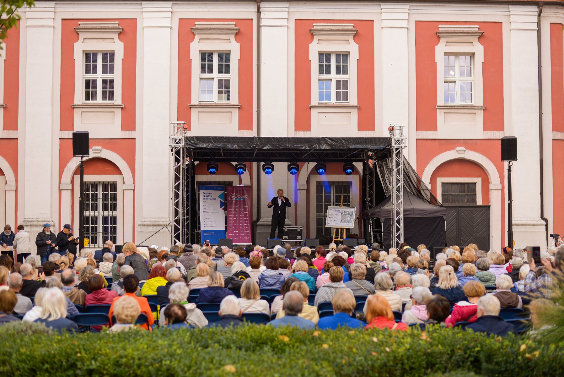 Na zdjęciu scena na dziedzińcu urzędu miasta, na scenie artysta, pod sceną słuchający go seniorzy
