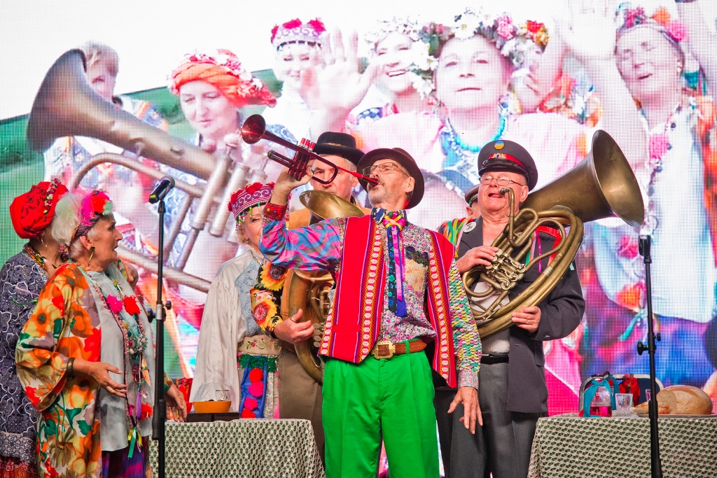 Na zdjęciu seniorzy w ludowych strojach na scenie, część z nich gra na instrumentach dętych