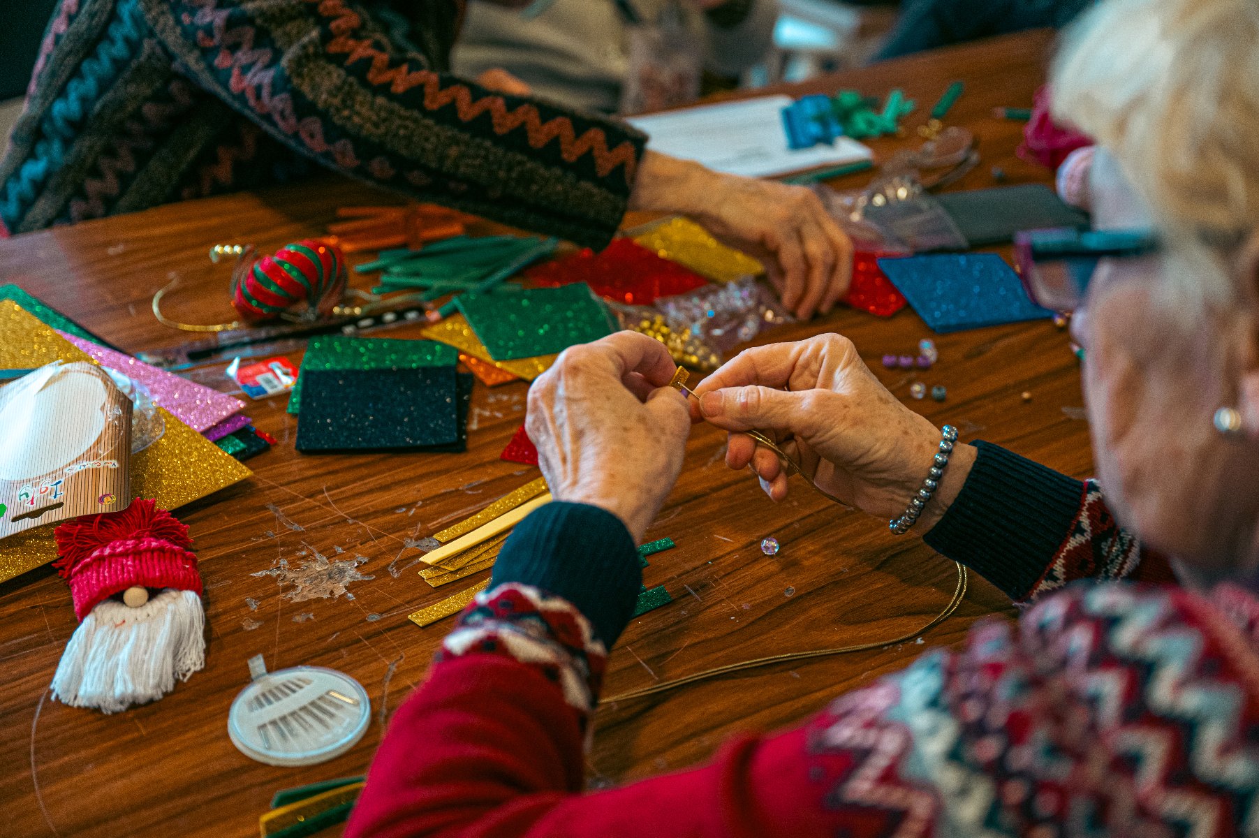 Na zdjęciu starsze kobiety robiące ozdoby choinkowe z kolorowych wstążek, koralików i papieru, widać tylko ich dłonie