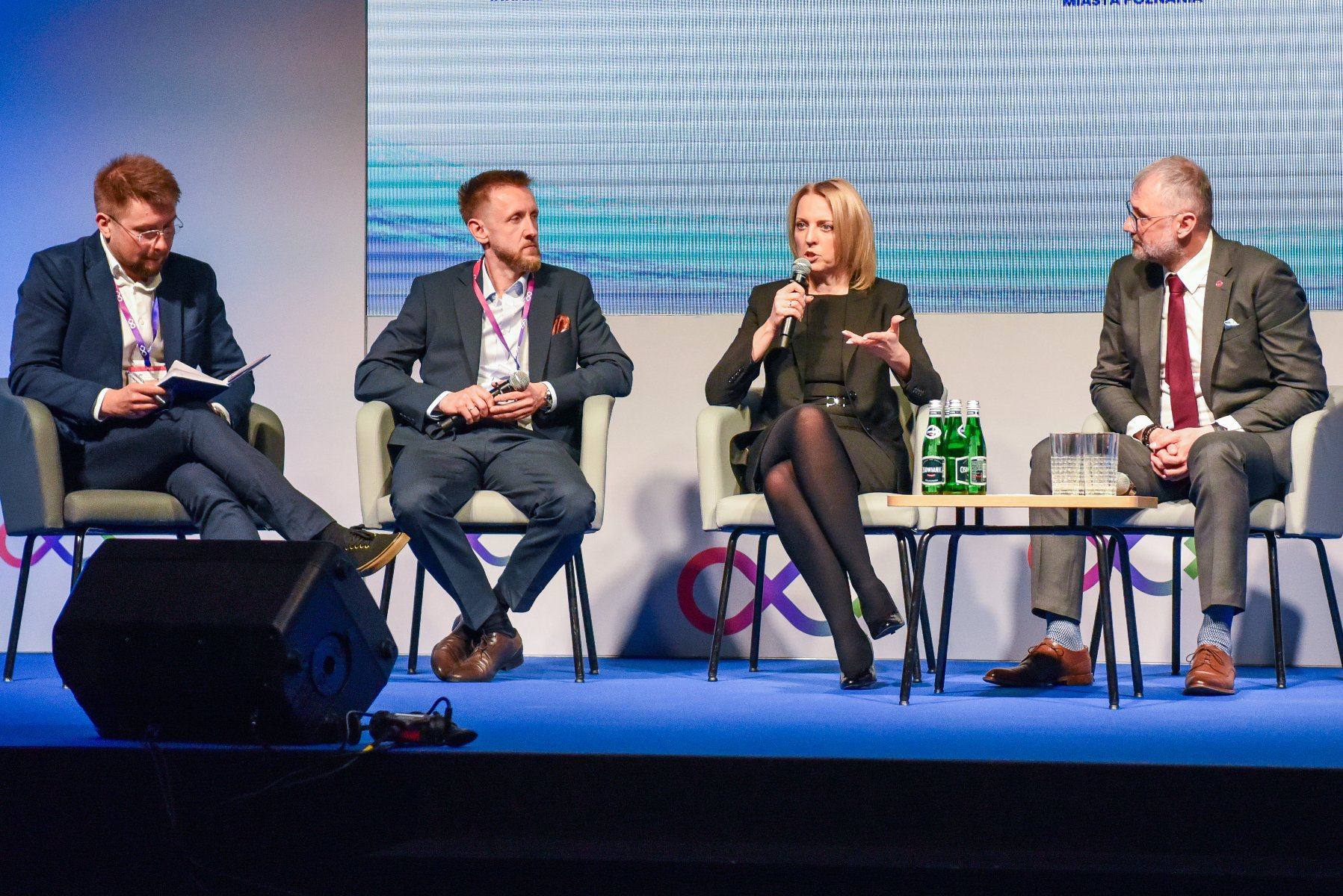 Na zdjęciu trzech mężczyzn i kobieta rozmawiają ze sobą, siedząc na krzesłach, kobieta trzyma mikrofon