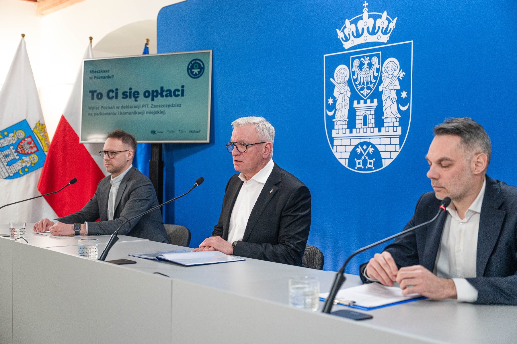 Na zdjęciu trzech mężczyzn za stołem konferencyjnym, w tle ścianka z herbem miasta