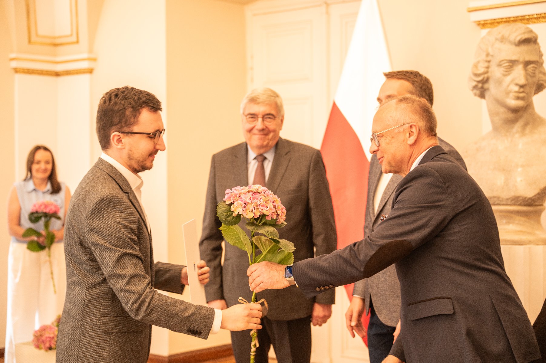 Na zdjęciu uroczystość wręczenia stypendiów. Trzech mężczyzn wręcza dyplom i kwiata.