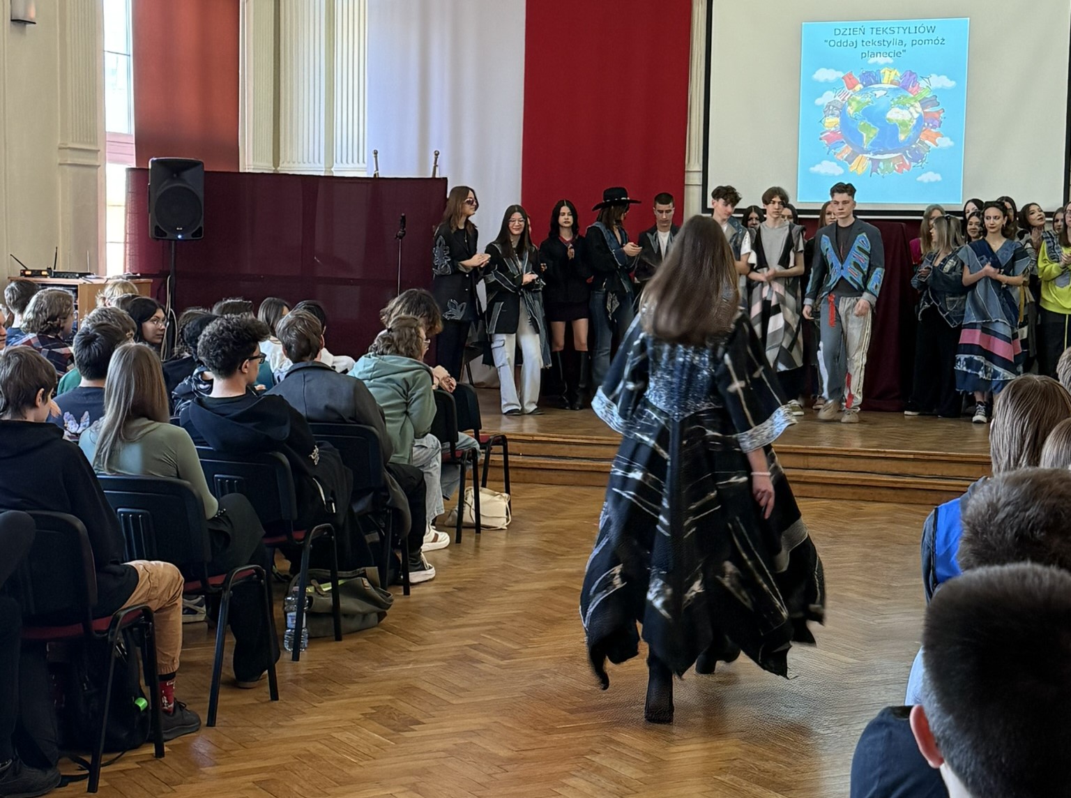 Na zdjęciu widać aulę szkolną, w której po lewej i prawej stronie siedzą widzowie, a na scenie z przodu i między krzesełkami odbywa się pokaz mody