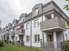 Na zdjęciu widać budynek, który ma 3 kondygnacje - parter i dwa piętra. Budynek widać od tyłu, nie od frontu. Przy każdym z mieszkań w tym budynku jest balkon. Przed budynkiem znajduje się trawnik.