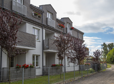 Na zdjęciu widać budynek w kolorystyce szaro-białej. Budynek ma parter i 2 piętra. Przed budynkiem jest teren zieleni, na którym rośnie trawa i drzewka. Budynek jest ogrodzony. Za ogrodzeniem jest droga.