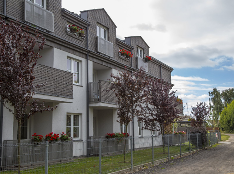 Na zdjęciu widać budynek w kolorystyce szaro-białej. Budynek ma parter i 2 piętra. Przed budynkiem jest teren zieleni, na którym rośnie trawa i drzewka. Budynek jest ogrodzony. Za ogrodzeniem jest droga.