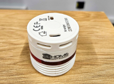 Na zdjęciu widać czujkę dymu. Czujka jest w kształcie walca, w białym kolorze, z napisem Home Safety Solution. Czujka stoi na drewnianym stole.