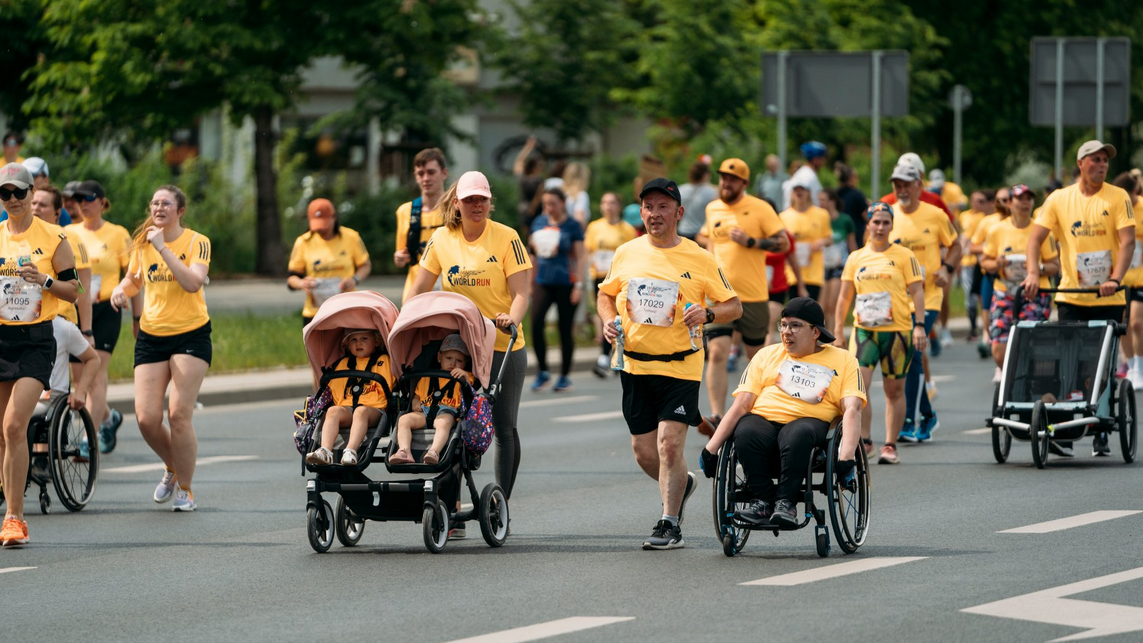 Na zdjęciu widać grupę osób w charakterystycznych żółtych koszulkach biegu Wings for Life, wśród których jest kobieta pchająca podwójny wózek z dziećmi, osoba na wózku inwalidzkim oraz inni biegacze poruszający się wspólnie szeroką jezdnią.