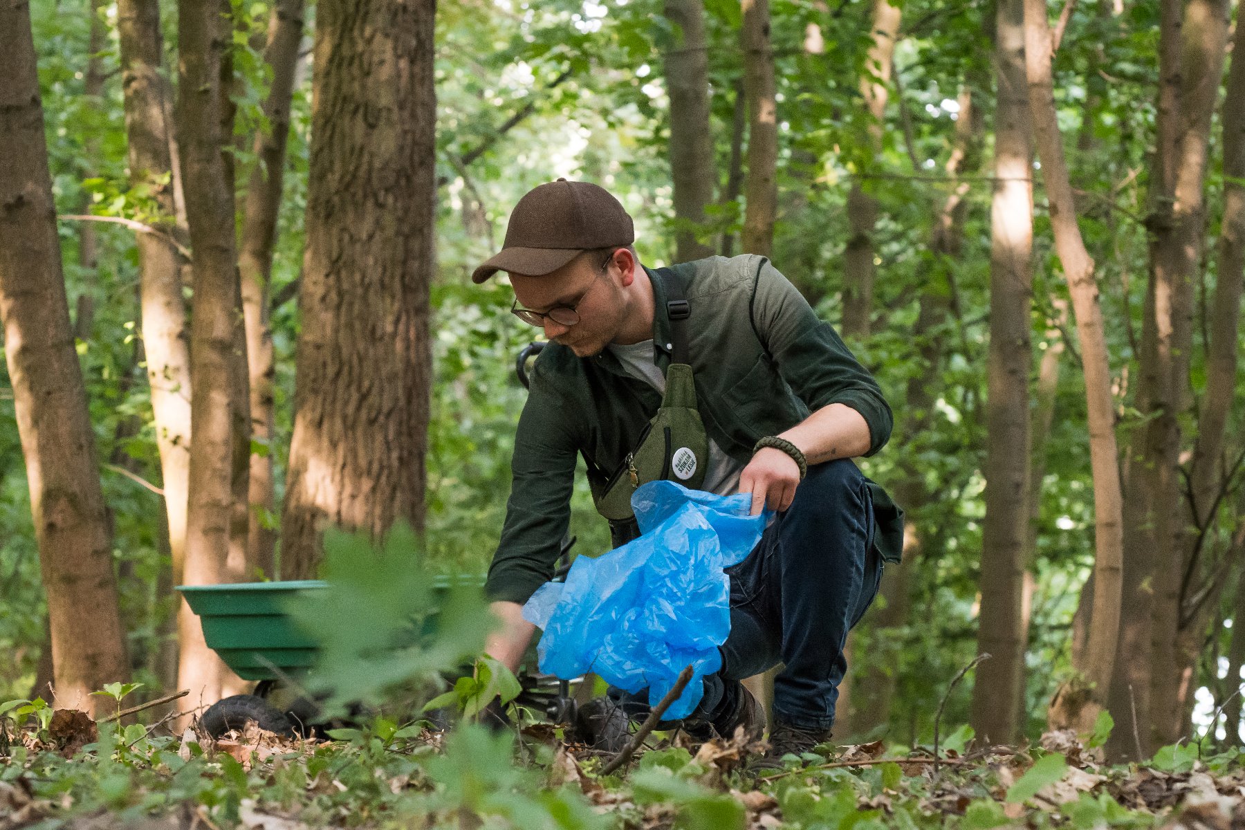 Na zdjęciu widać mężczyznę w lesie, któy przykucnął z workiem na śmieci