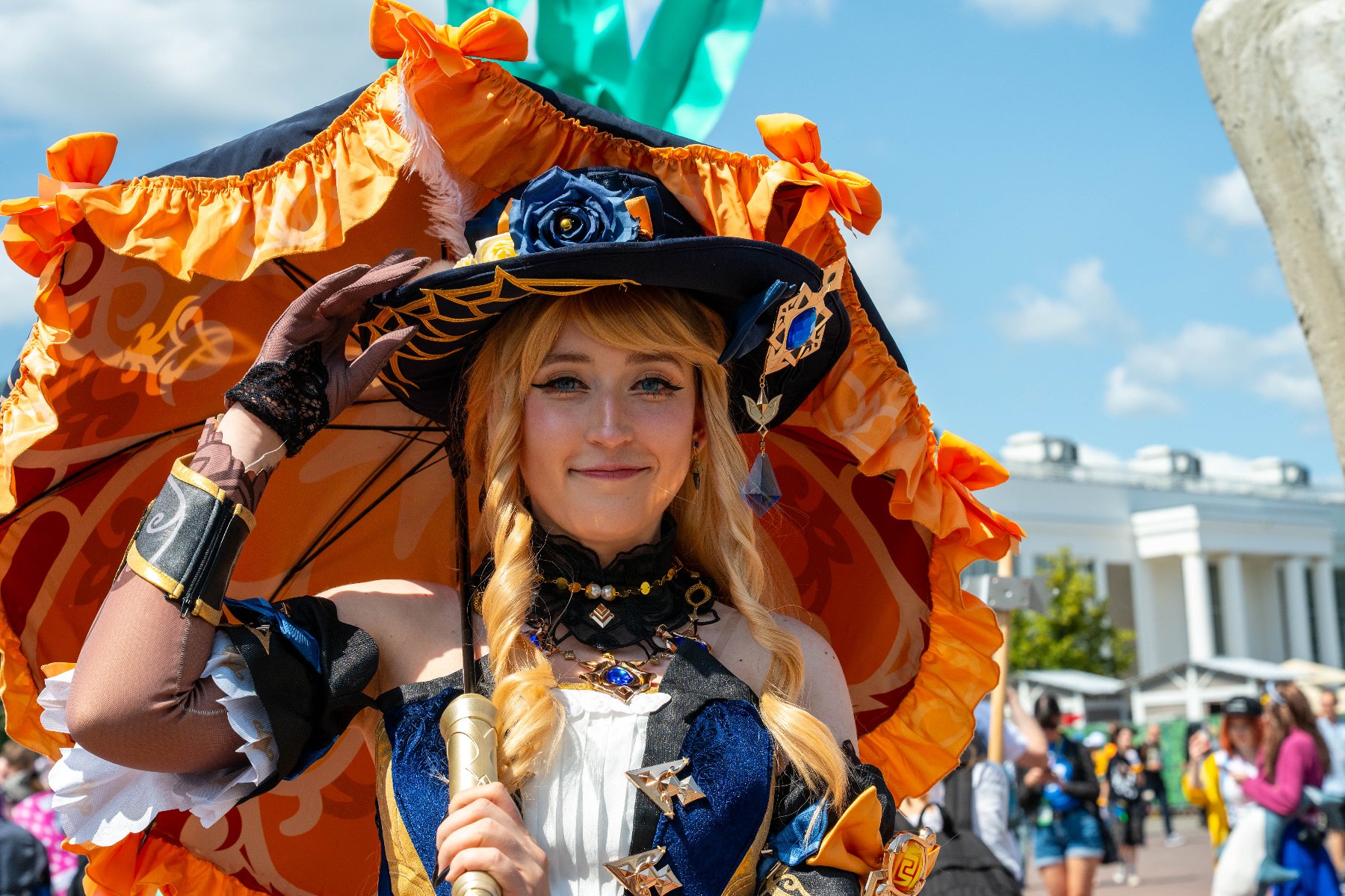 Na zdjęciu widać młodą kobietę w kolorowym cosplayu, przebrana za postać o bogatym, eleganckim stroju - nosi kapelusz zdobiony niebieskimi kwiatami i pomarańczową parasolkę z ozdobnymi falbanami. W tle widać tłum ludzi oraz biały budynek, a cała scena rozgrywa się w słoneczny, letni dzień na otwartym powietrzu.