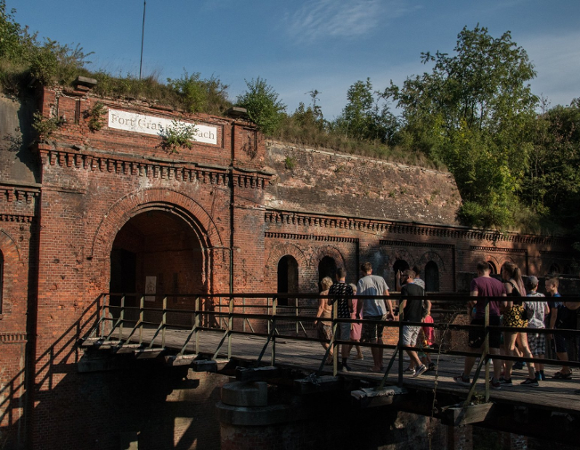 Na zdjęciu widać monumentalną budowlę z czerwonej cegły z charakterystycznymi łukowatymi sklepieniami i otworami strzelniczymi. Nad głównym wejściem widnieje tablica z napisem. Przez fosę prowadzi drewniany most, po którym idzie grupa zwiedzających. Obiekt jest częściowo porośnięty roślinnością, co podkreśla jego historyczny, surowy charakter. Całość została uchwycona w świetle dziennym, co uwydatnia detale starej architektury militarnej.