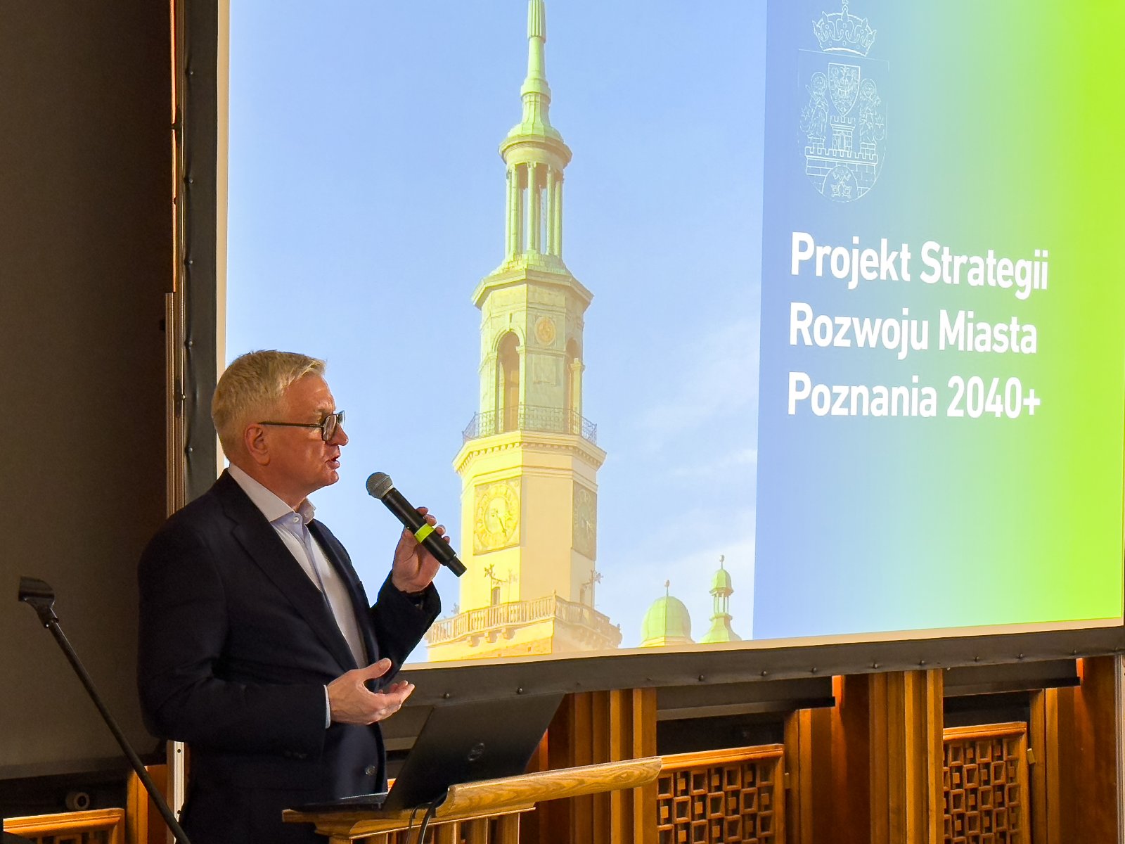 na zdjęciu widac pana prezydenta miasta Poznania Jacka Jaśkowiaka, który w ręce trzyma mikrofon i przemawia. Za nim widać prezentację wyświetalną na projektorze, na której widać ratusz poznański oraz napis "Projekt Strategii Rozwoju Miasta Poznania 2040+"