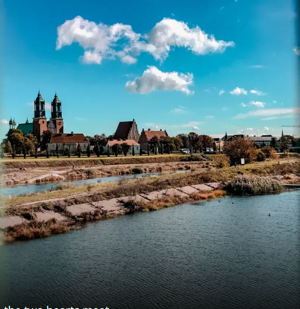 Na zdjęciu widać panoramę miasta nad rzeką, z charakterystycznymi zabudowaniami i wieżami kościoła górującymi nad okolicą. Woda, nadrzeczne tereny i spokojny krajobraz tworzą malowniczy widok w słoneczny dzień.