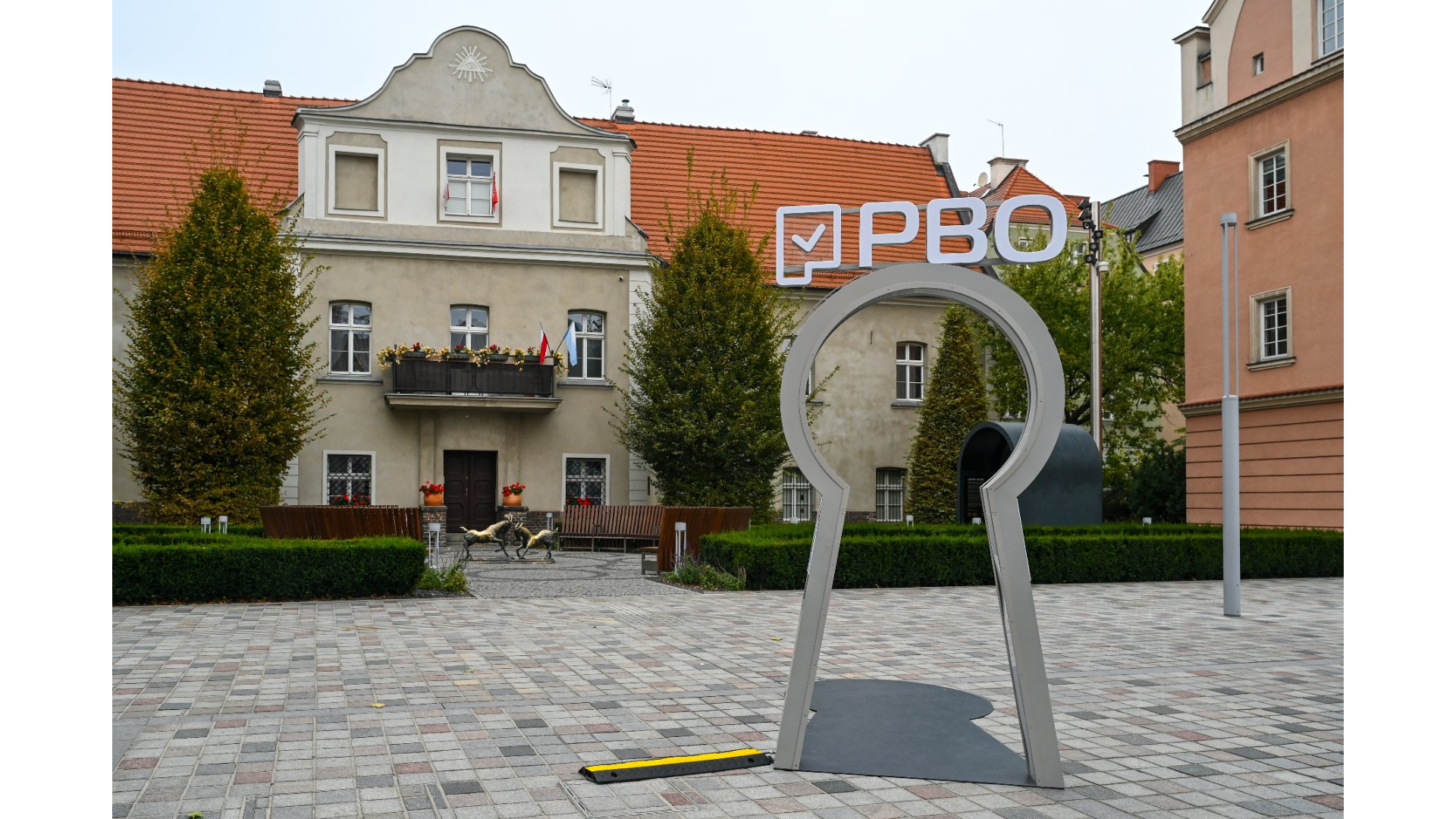 Na zdjęciu widać rzeźbę symbol Poznańskiego Budżetu Obywatelskiego, stojącą na kostce brukowej, za nią w tle budynek, przed którym stoją figurki dwóch poznańskich koziołków.