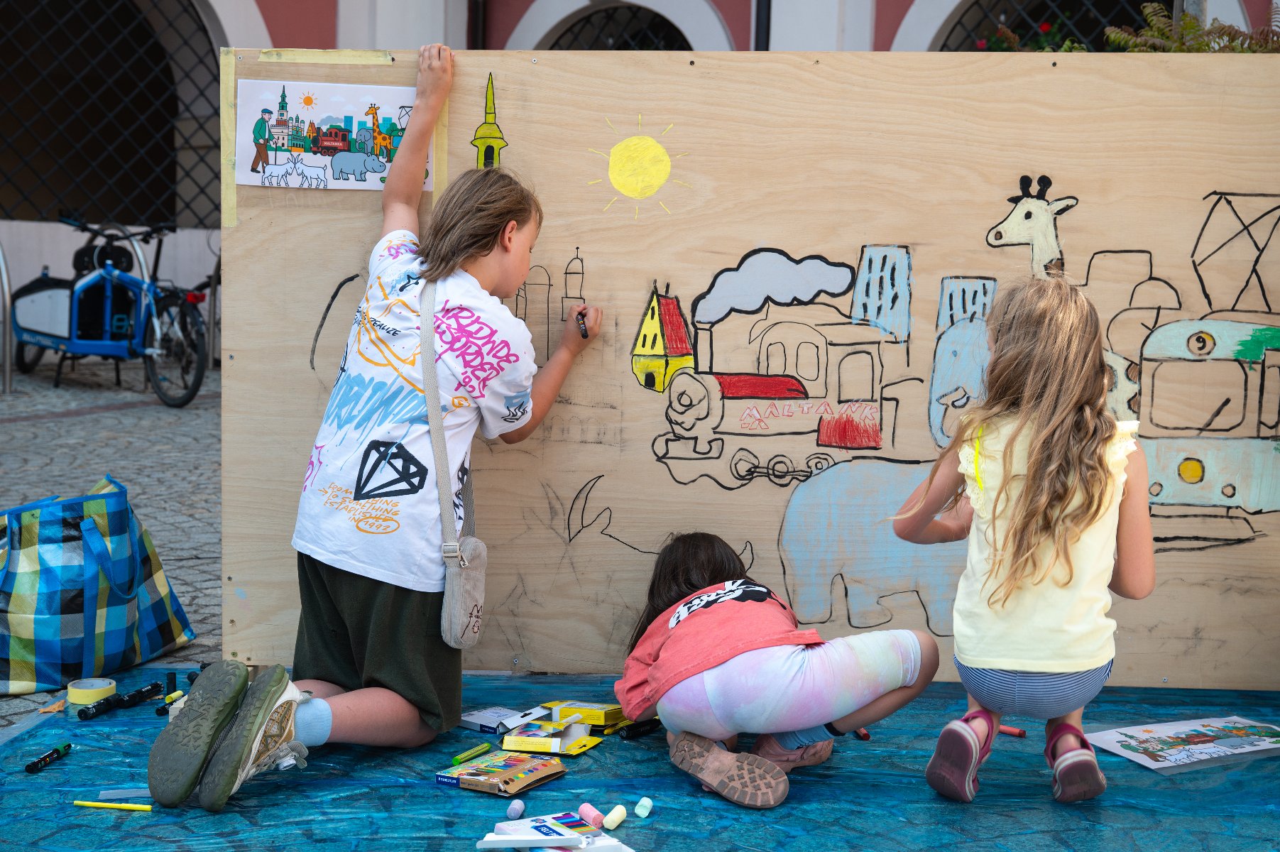 Na zdjęciu widać troje dzieci rysujących i kolorujących duży mural na drewnianej ścianie. Na ilustracji znajdują się słoń, żyrafa, nosorożec, tramwaj, lokomotywa z napisem "MALTANKA", budynki oraz słońce. Obok po lewej stronie stoją rowery i torba.