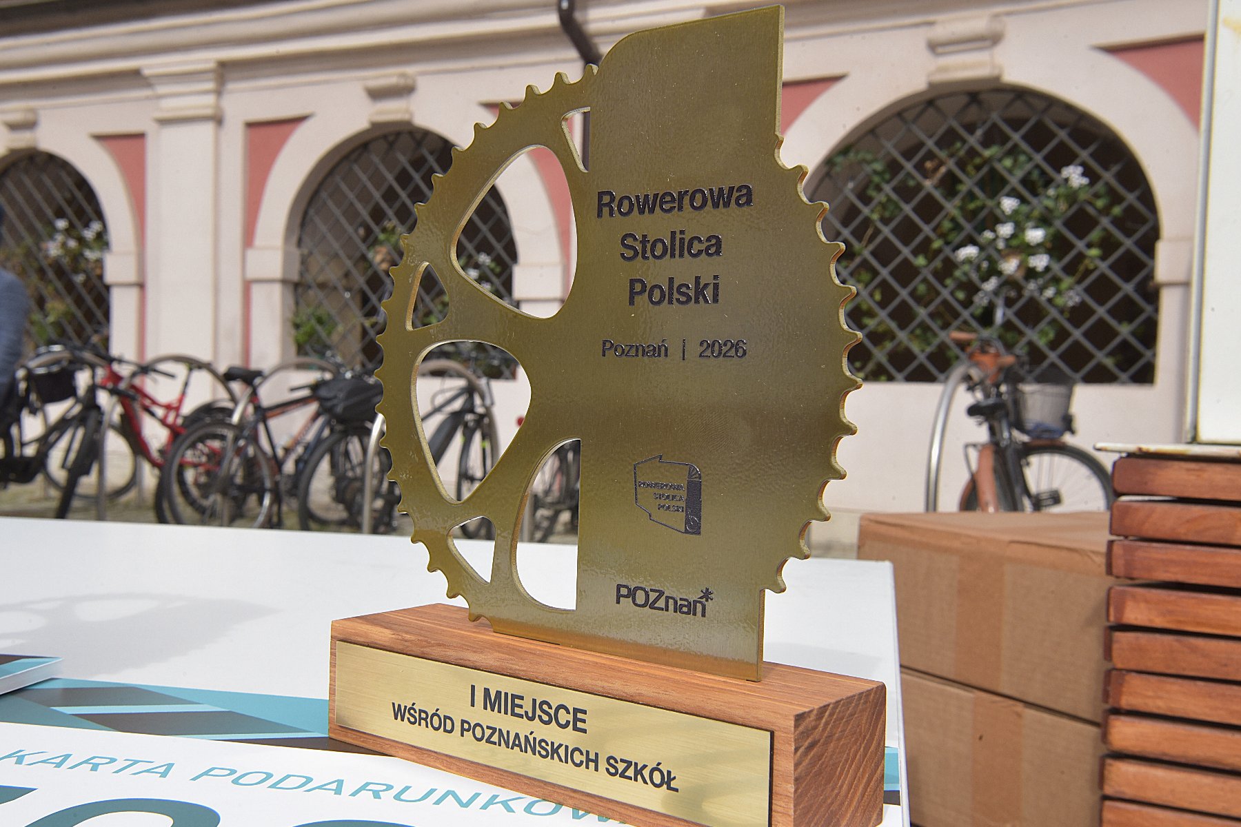 Na zdjęciu widoczna jest statuetka w kształcie zębatki rowerowej z napisem "Rowerowa Stolica Polski, Poznań 2026", ustawiona na drewnianej podstawie z tabliczką "I miejsce wśród poznańskich szkół". W tle znajdują się zaparkowane rowery oraz arkadowa elewacja budynku.