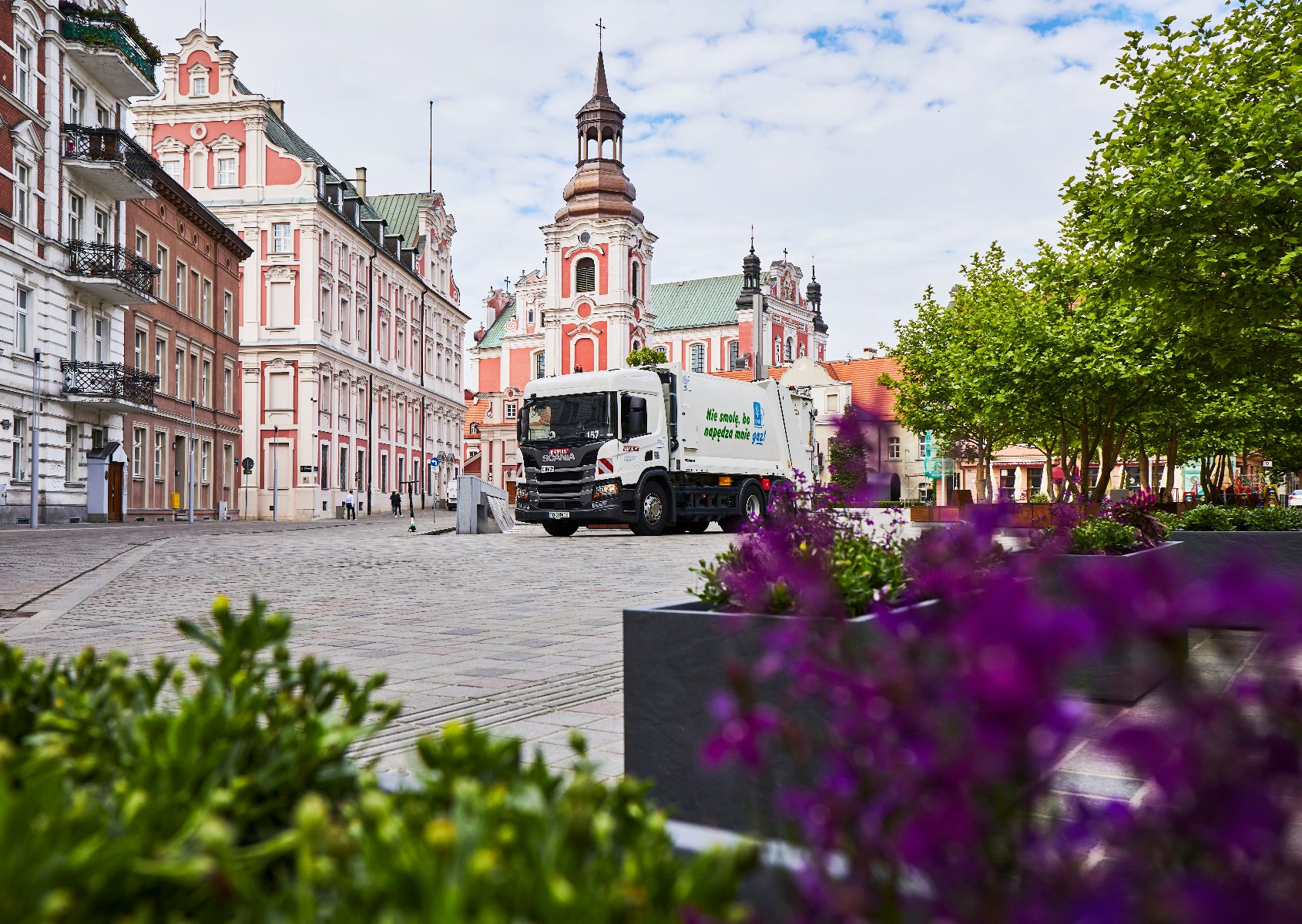 Na zdjęciu znajdują się kwiaty oraz budynek Urzędu Miasta Poznania i kościoła farnego. Na środku również stoi śmieciarka.
