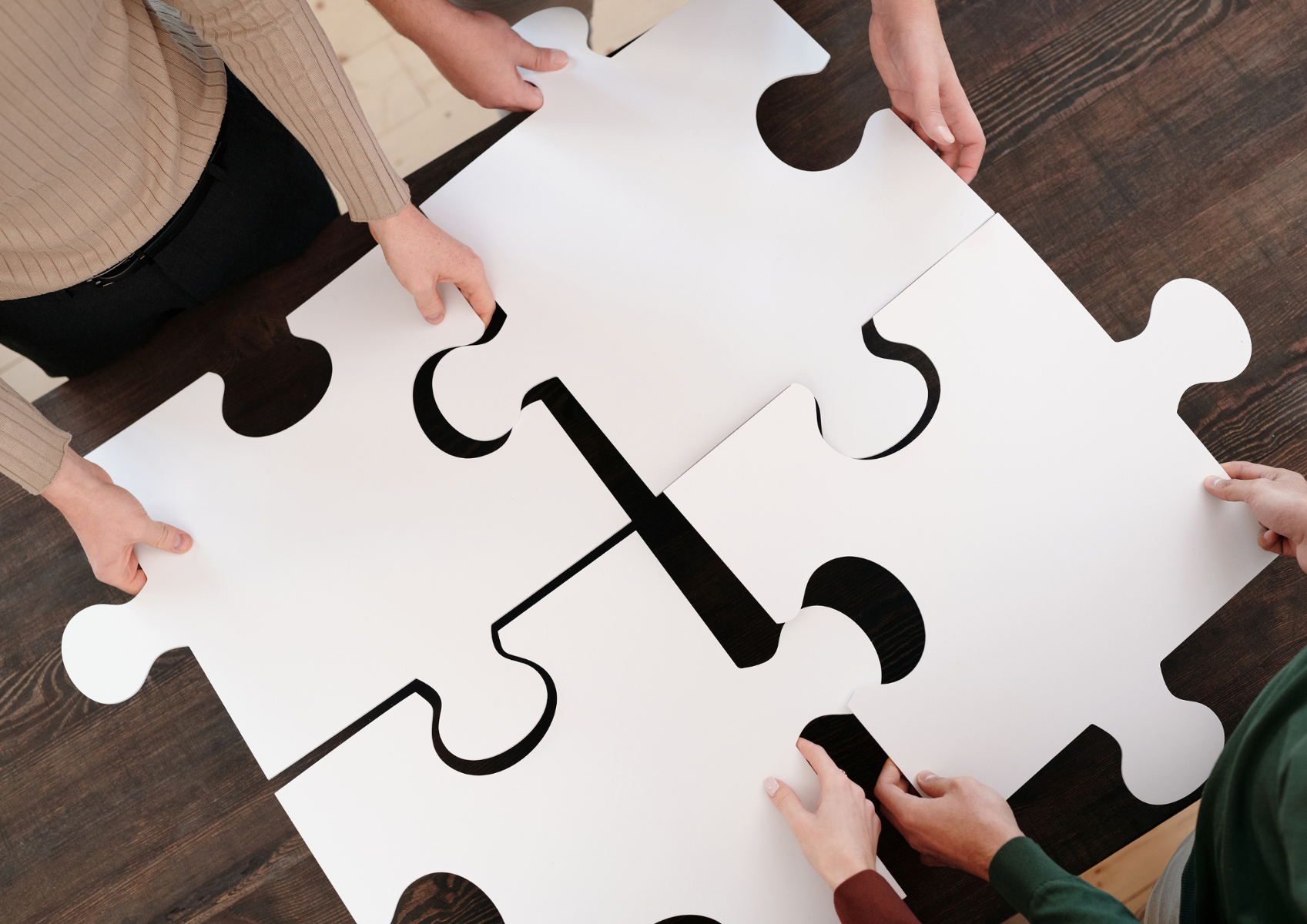 Na zdjęciu znajdują się ręce 4 osób trzymające duże kawałki białych puzzli tak, aby połączyły się ze sobą.