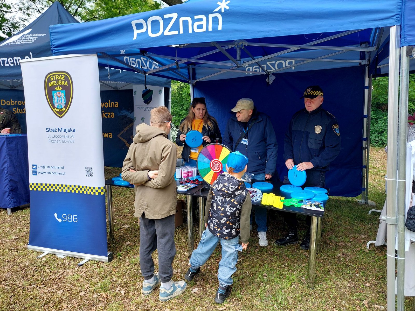 Na zdjęciu znajduje się niebieski namiot z logo Poznania, 3 osoby ze straży miejskiej, 2 kobiety i mężczyzna prezentują materiały promocyjne przy stole, na stole jest kolorowe koło do kręcenia i losowania zadań, przed namiotem stoi dwóch chłopców