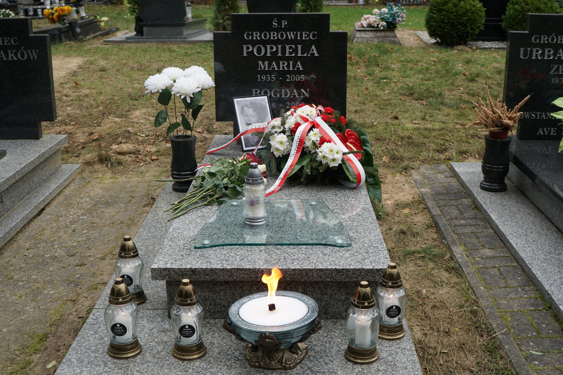 Nagrobek rodziny Popiela, w której spoczywa Maria i Bogdan. Nagrobek jest przystrojony wieńcem i zdjęciem Marii Popieli. Przed narobkiem pali się symboliczne światełko dla Czerwca '56. Wokół stoją zapalone znicze.