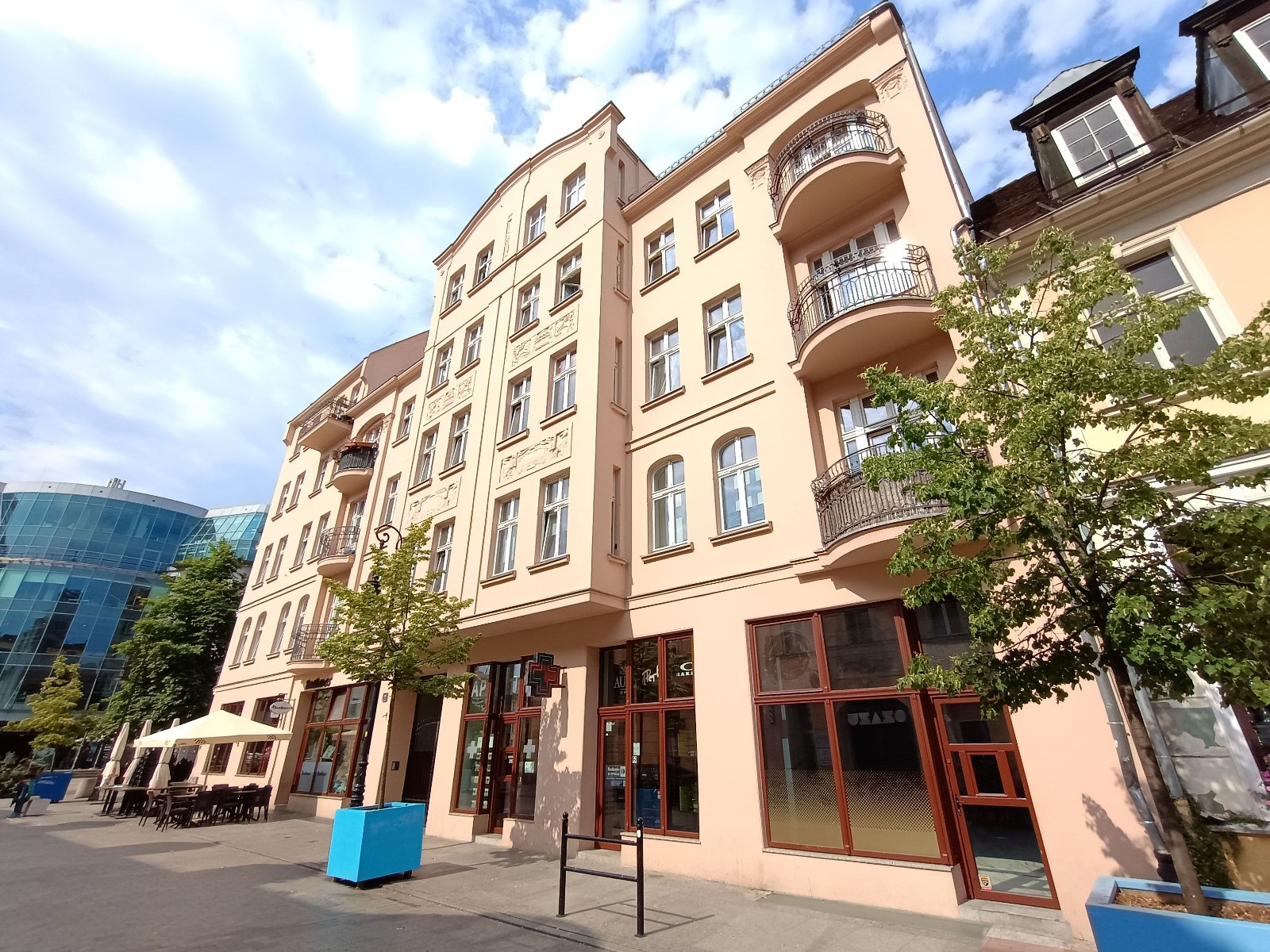 Narożny budynek mieszkalno-usługowy o jasnej, beżowej fasadzie, z rzędami okien oraz balkonami z metalowymi balustradami, położony przy ulicy z dużymi drzewami.