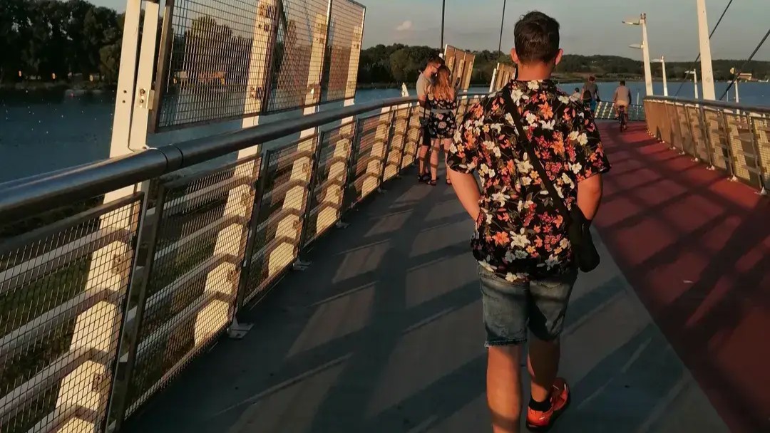 Nastolatek idzie po moście nad wodą o zachodzie słońca. W tle widoczni są spacerowicze, rowerzyści oraz zieleń wokół jeziora.