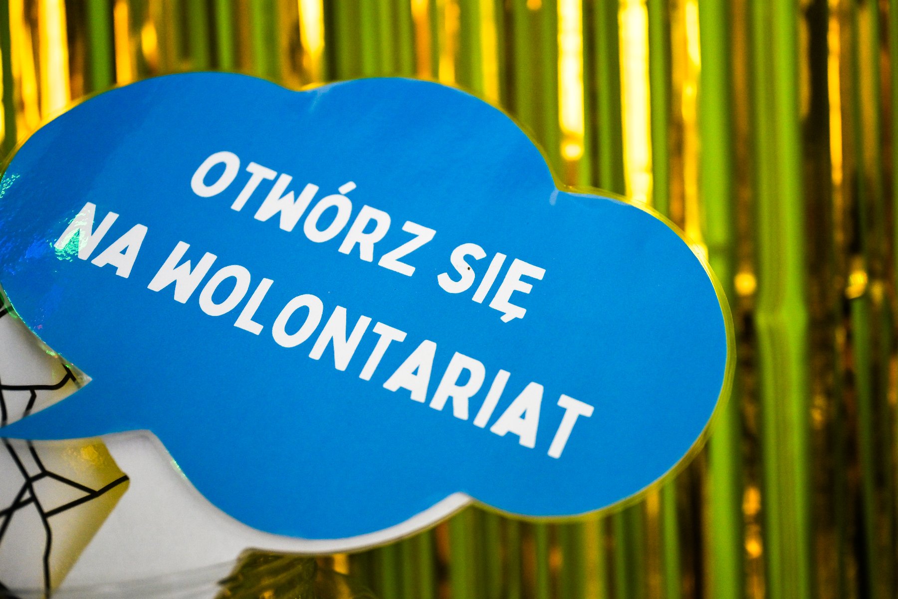 Niebieska tabliczka w kształcie chmurki dialogowej z napisem "Otwórz się na wolontariat" na tle ozdobnej kurtyny.