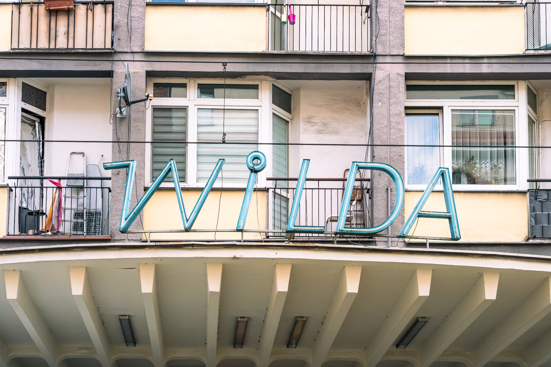 Niebieski, retro neon z napisem "WILDA" zamontowany na elewacji budynku mieszkalnego, tuż nad zadaszeniem z charakterystycznymi filarami.
