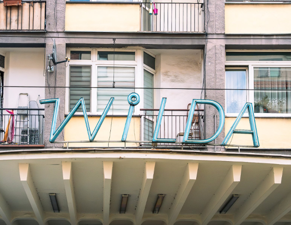 Niebieski, retro neon z napisem "WILDA" zamontowany na elewacji budynku mieszkalnego, tuż nad zadaszeniem z charakterystycznymi filarami.