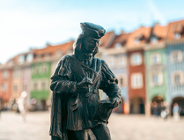 Niewielka figurka z brązu przedstawiająca postać w historycznym stroju z rysownikiem lub księgą, ustawiona na tle rozmytych, kolorowych kamieniczek rynkowych.