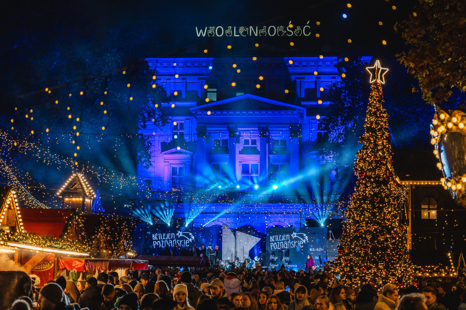 Noc. Widok na podświetlony na niebiesko budynek Arkadii na Placu Wolności. Nad budynkiem napis "WOLNOŚĆ". Przed budynkiem rozświetlone Betlejem Poznańskie, czyli budki i wysoka choinka. Środkiem chodzi tłum ludzi.