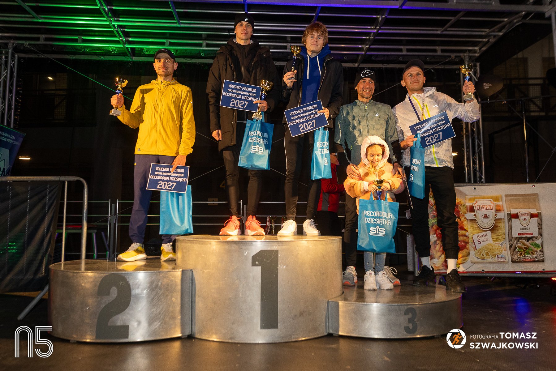 Nocna Recordowa Piątka 2026, fot. Tomasz Szwajkowski - na podium 5 mężczyzn i 1 dziecko. Wszyscy trzymają nagrody i vouchery i puchary