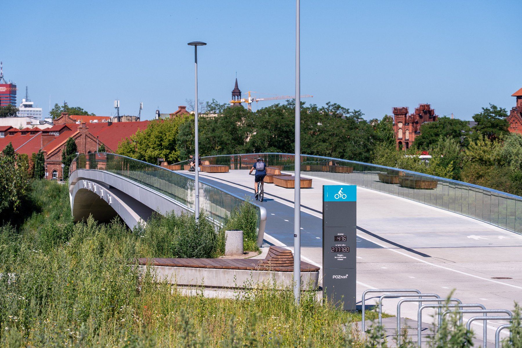 Nowoczesna kładka pieszo-rowerowa nad Wartą w Poznaniu, z widocznym rowerzystą jadącym po jasnej ścieżce rowerowej. Obok ścieżki znajduje się ciemnoszary słupek z niebieskim piktogramem roweru i licznikiem rowerowym. W tle widać zabudowania Poznania, w tym czerwone ceglane budynki i dźwig.
