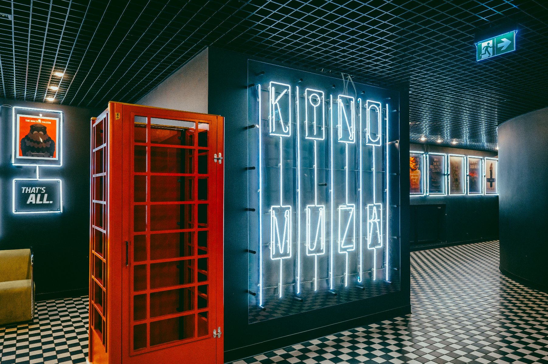 Nowoczesne foyer kina: Czerwona budka telefoniczna obok dużej, podświetlanej na niebiesko instalacji neonowej z napisem "KINO MUZA". Na ścianach plakaty w podświetlanych ramach. Podłoga w czarno-białą szachownicę, sufit z czarnej siatki