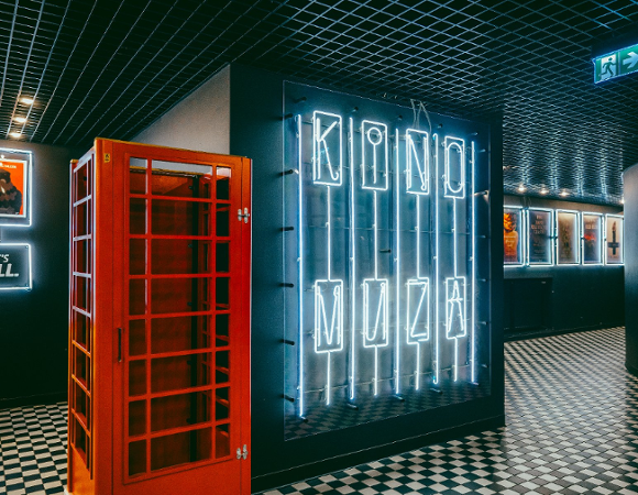 Nowoczesne foyer kina: Czerwona budka telefoniczna obok dużej, podświetlanej na niebiesko instalacji neonowej z napisem "KINO MUZA". Na ścianach plakaty w podświetlanych ramach. Podłoga w czarno-białą szachownicę, sufit z czarnej siatki