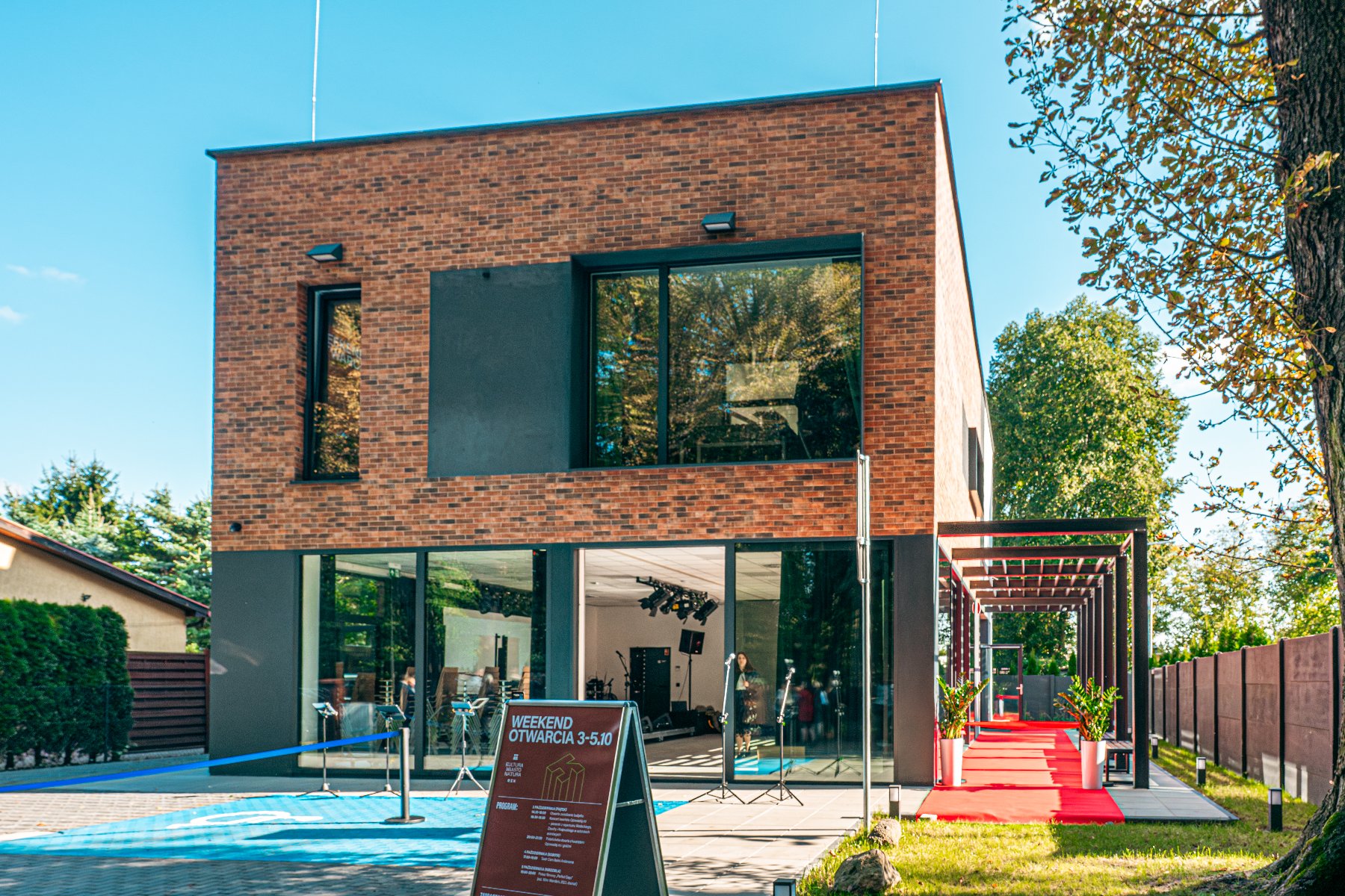 nowoczesny, dwupiętrowy budynek o elewacji z ciemnej cegły i dużymi, panoramicznymi oknami, otwarty na czerwony dywan prowadzący na zewnątrz. Przed wejściem stoi tablica informacyjna z napisem "WEEKEND OTWARCIA 3-5.10", a budynek jest otoczony zielenią