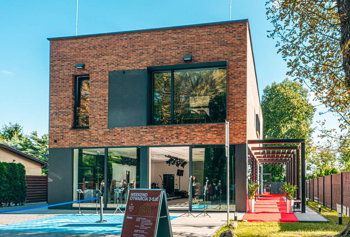 nowoczesny, dwupiętrowy budynek o elewacji z ciemnej cegły i dużymi, panoramicznymi oknami, otwarty na czerwony dywan prowadzący na zewnątrz. Przed wejściem stoi tablica informacyjna z napisem "WEEKEND OTWARCIA 3-5.10", a budynek jest otoczony zielenią - grafika artykułu