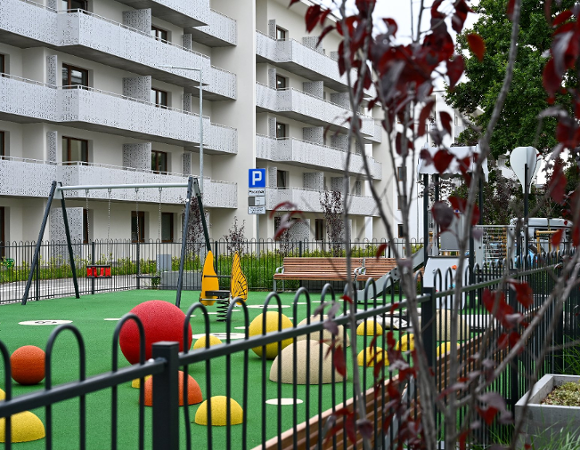 Nowoczesny plac zabaw z zieloną nawierzchnią na osiedlu. Na pierwszym planie czarne metalowe ogrodzenie i gałęzie z czerwonymi liśćmi. Na placu kolorowe półkule, huśtawki oraz żółta zabawka na sprężynie w kształcie motyla. W tle biały wielopiętrowy budynek z licznymi balkonami.