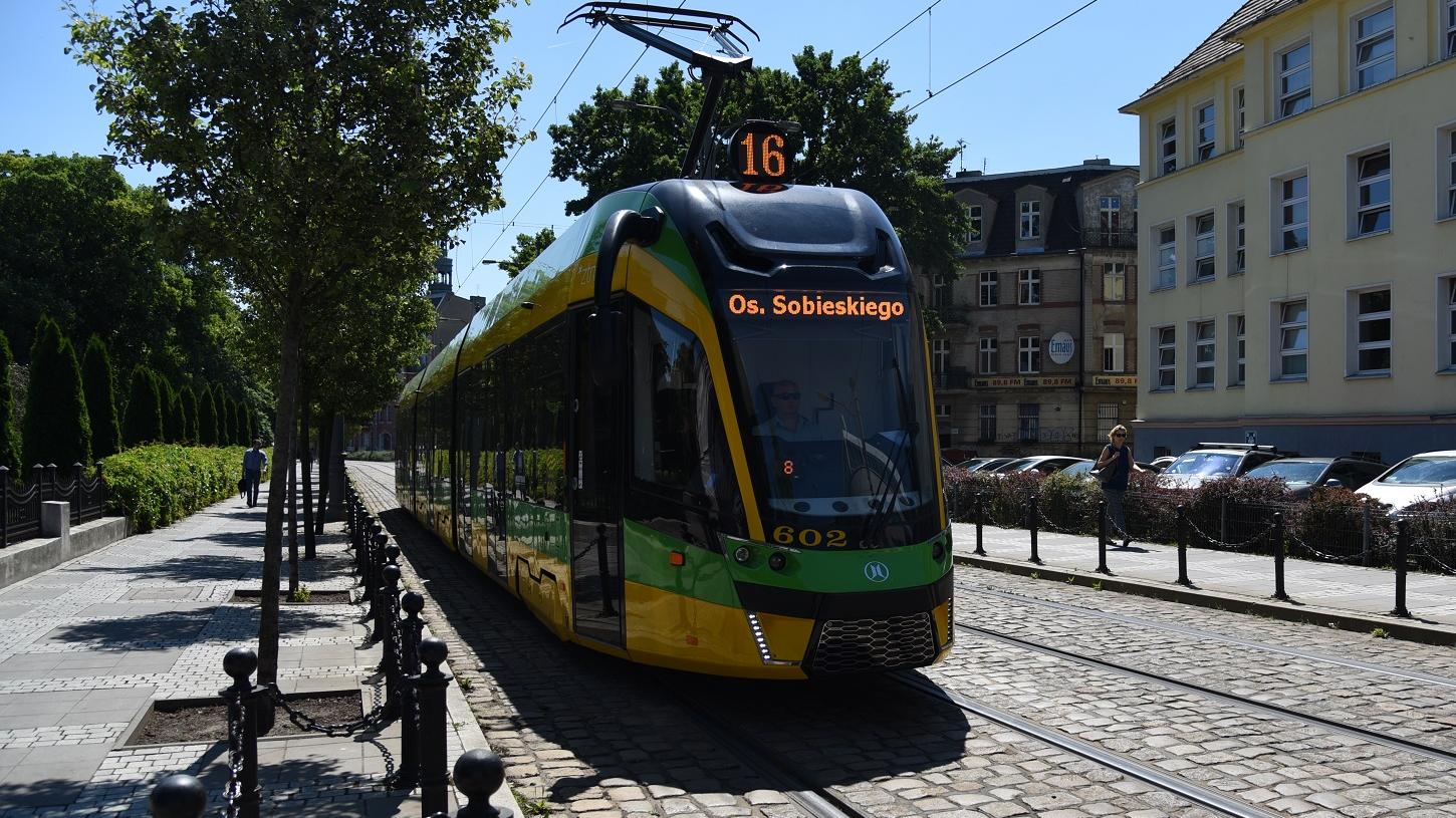 Nowoczesny tramwaj linii 16 jadący ulicą w słoneczny dzień, otoczony drzewami i zabudową miejską.