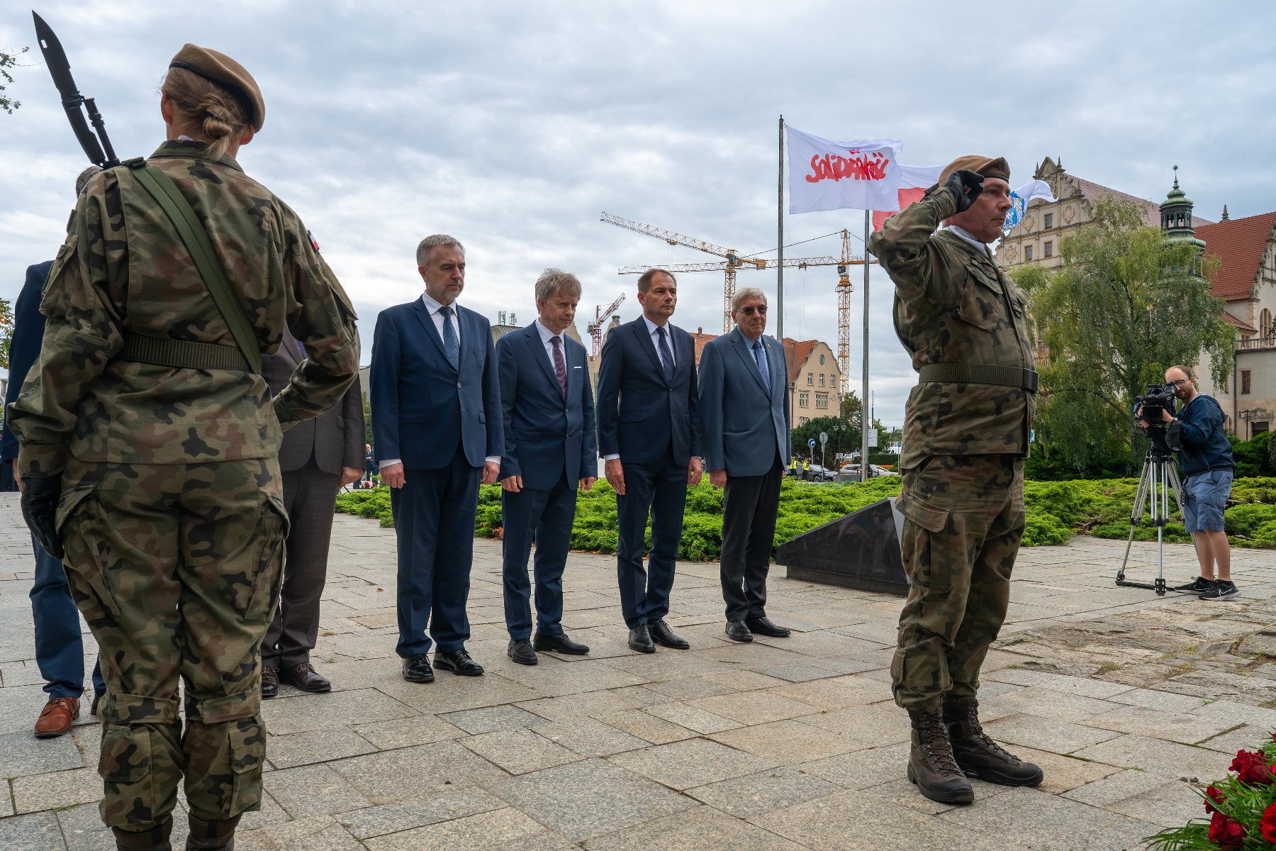Oficjalna delegacja mężczyzn w garniturach składająca kwiaty podczas uroczystości patriotycznej w obecności wojskowej warty honorowej.