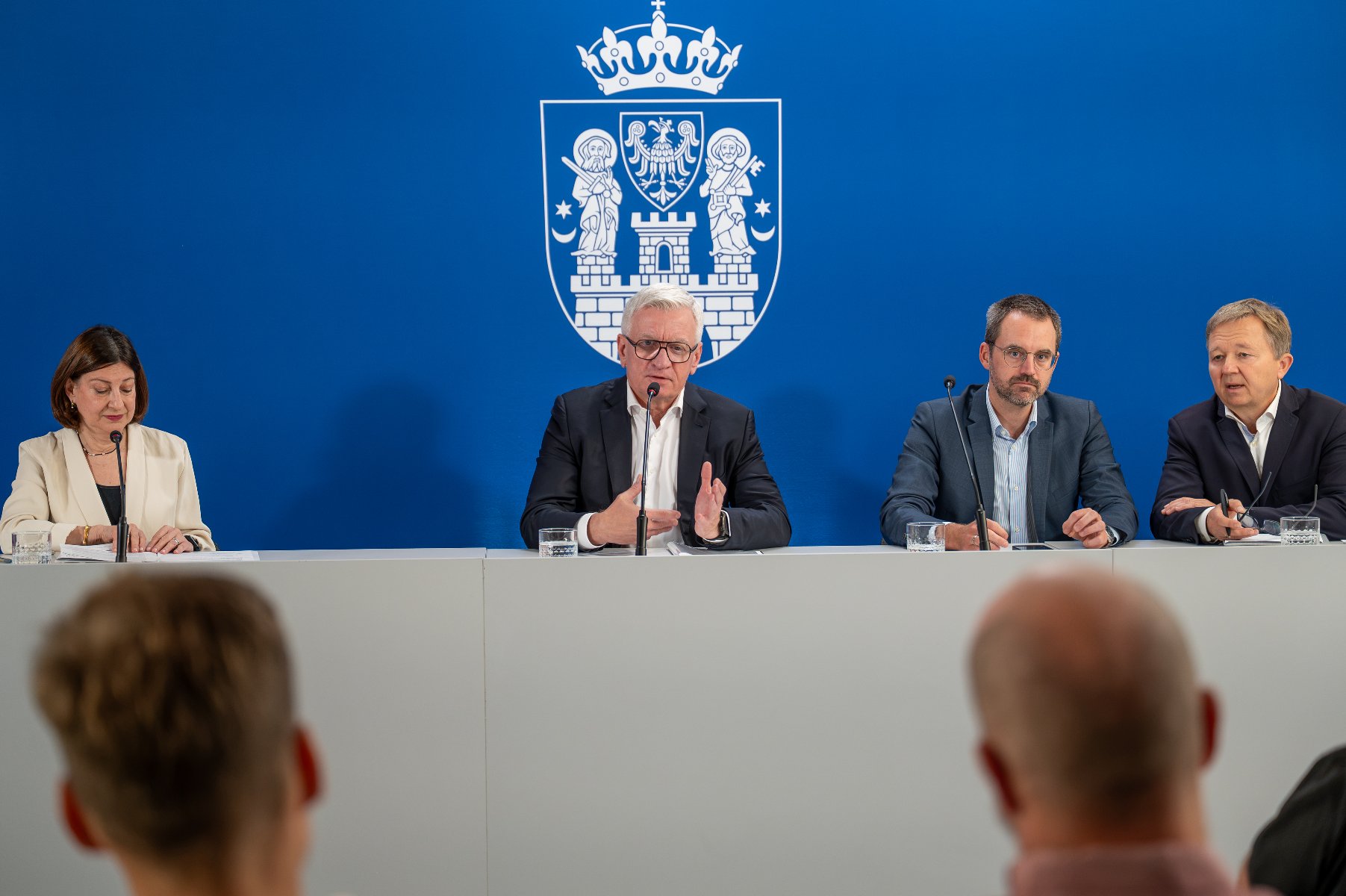 Oficjalna konferencja prasowa władz miasta Poznań z udziałem prezydenta Jacka Jaśkowiaka na tle niebieskiego ścianki z herbem miasta.