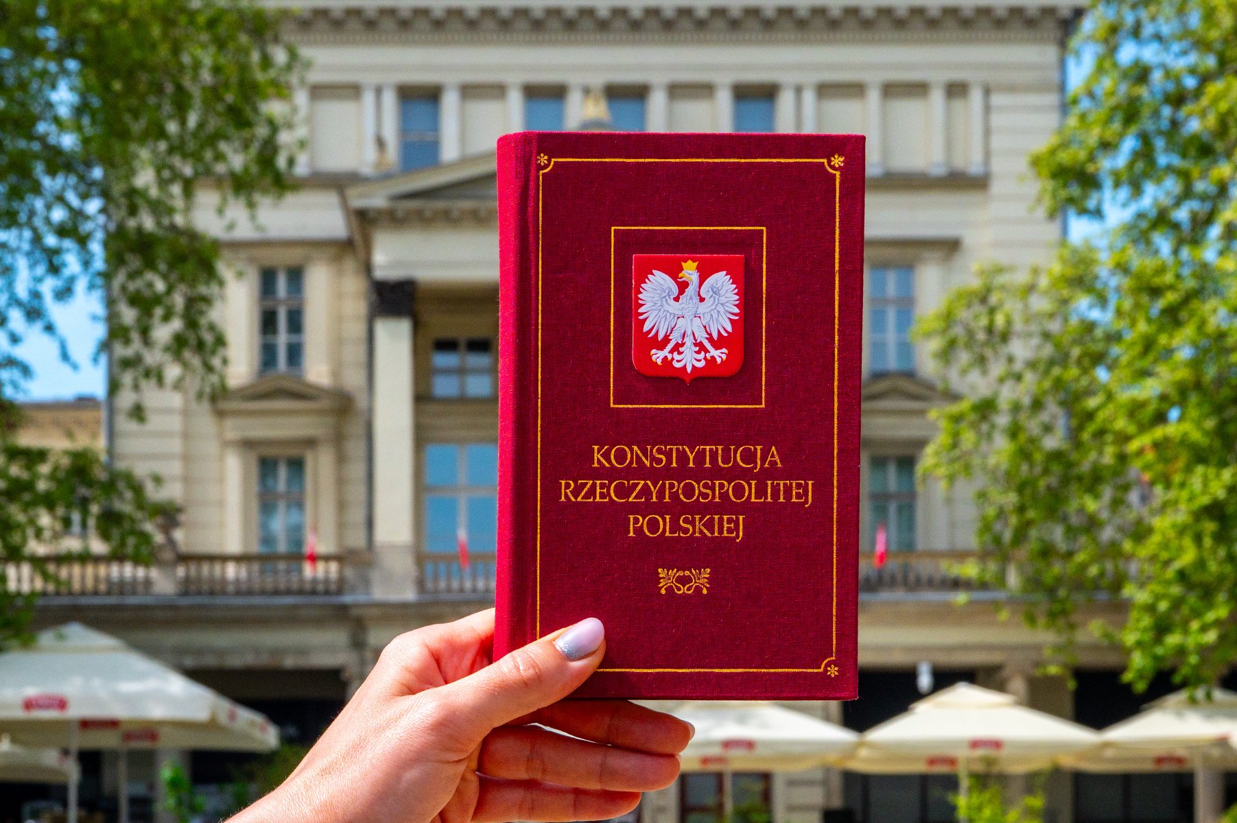 Osoba trzyma egzemplarz Konstytucji Rzeczypospolitej Polskiej. W tle budynek Arkadii przy Placu Wolności