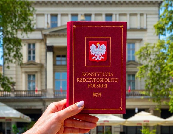 Osoba trzyma egzemplarz Konstytucji Rzeczypospolitej Polskiej. W tle budynek Arkadii przy Placu Wolności
